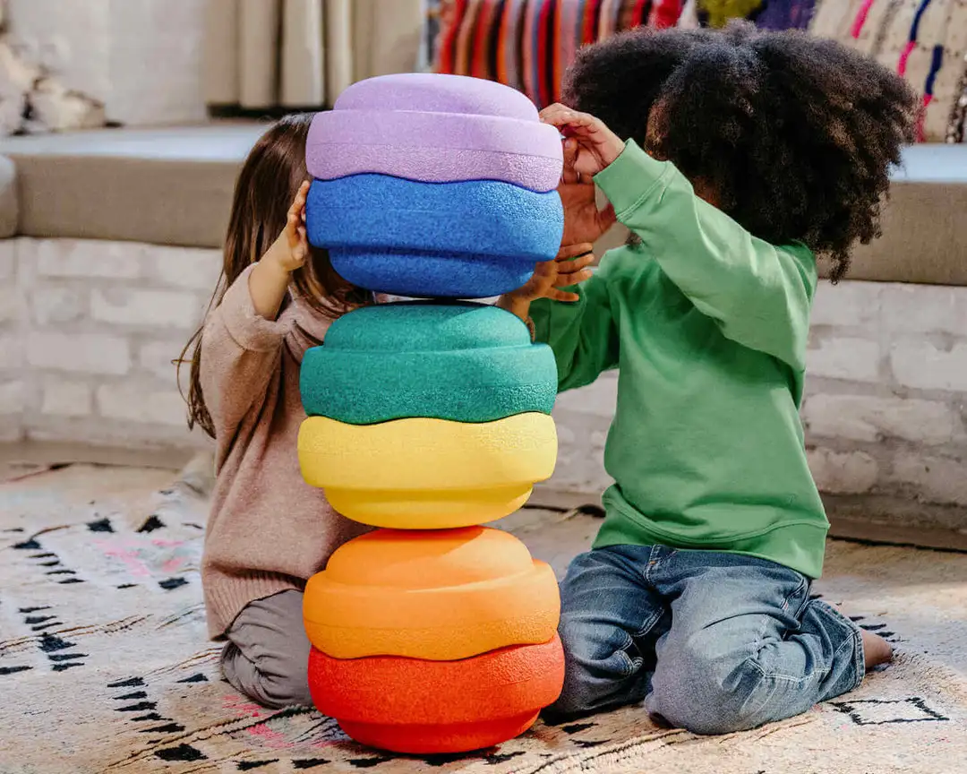 Zwei Kinder stapeln bunte Stapelstein Rainbow Elemente und spielen gemeinsam auf dem Teppich